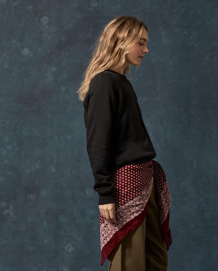 A person with long, light hair stands in profile against a blue textured background, wearing THE GREAT.’s The Fleece Rookie Sweatshirt in solid Almost Black, paired with brown pants and a red patterned scarf tied at the waist—an effortlessly cool look inspired by vintage classics made in Los Angeles.