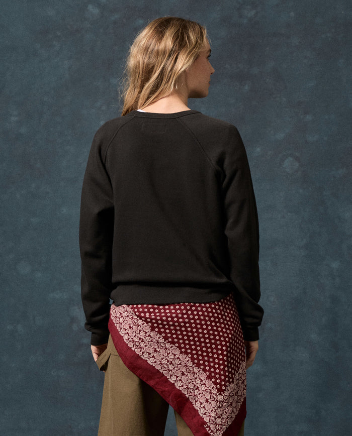 A person with long, light brown hair faces away, wearing THE GREAT. F26 D1 FLEECE BACK KNITS The Fleece Rookie Sweatshirt in Solid -- Almost Black, paired with khaki pants and a maroon bandana with a white pattern tied around the waist, set against a blue textured backdrop. This vintage sweatshirt is made in Los Angeles.