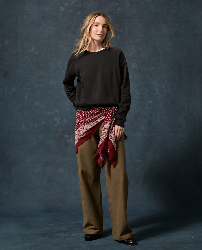 A person stands against a dark textured backdrop wearing THE GREAT.’s The Fleece Rookie Sweatshirt in Solid -- Almost Black, paired with loose brown pants, black shoes, and a red-patterned scarf tied around the waist.