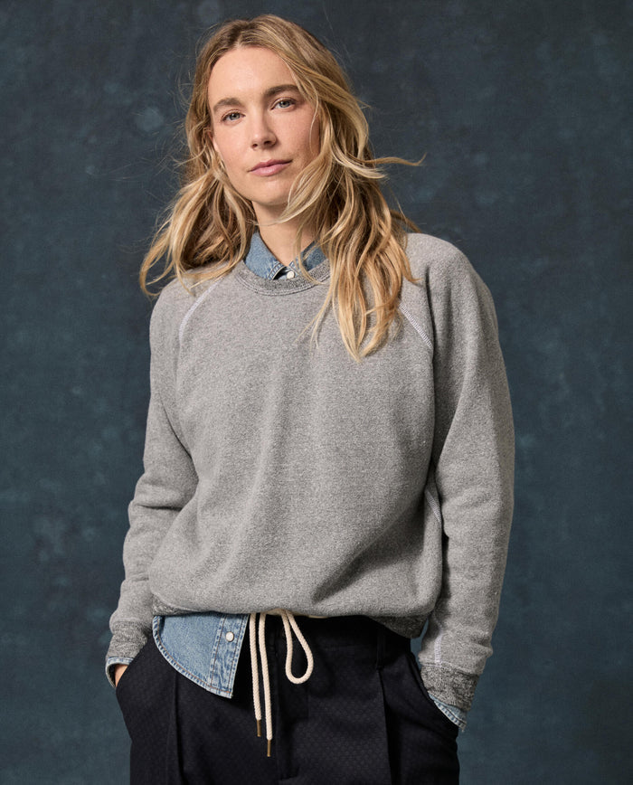 A person with long blond hair wears THE GREAT.’s The Fleece Rookie Sweatshirt in solid dark heather grey over a denim shirt and dark pants, standing against a dark mottled background.