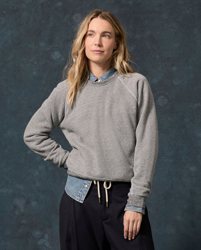 A woman with long blonde hair stands against a dark mottled background, wearing THE GREAT. F26 D1 FLEECE BACK KNITS “The Fleece Rookie Sweatshirt” in Solid Dark Heather Grey over a buttoned denim shirt and black pants.