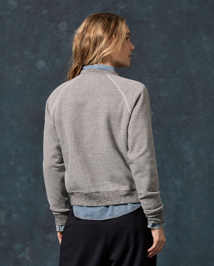 A person with long hair, dressed in THE GREAT. F26 D1 FLEECE BACK KNITS The Fleece Rookie Sweatshirt, Solid -- Dark Heather Grey, layered over a denim shirt and paired with black pants, stands facing away against a dark textured background.