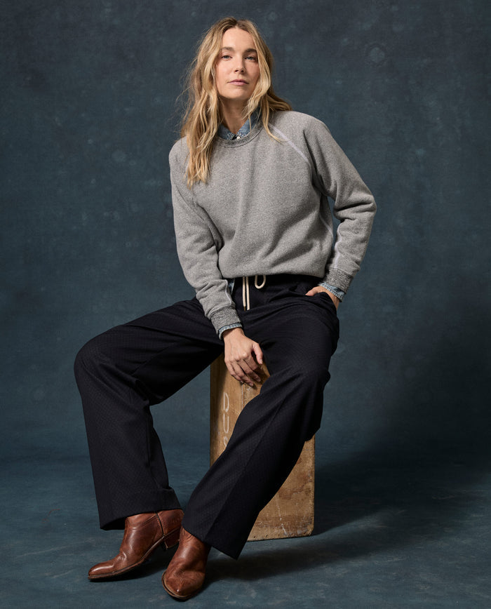 A person with long blonde hair sits on a wooden block, wearing THE GREAT.’s Fleece Rookie Sweatshirt in Solid Dark Heather Grey from the F26 D1 FLEECE BACK KNITS line over a blue shirt, paired with wide black pants and brown boots, set against a dark mottled backdrop.