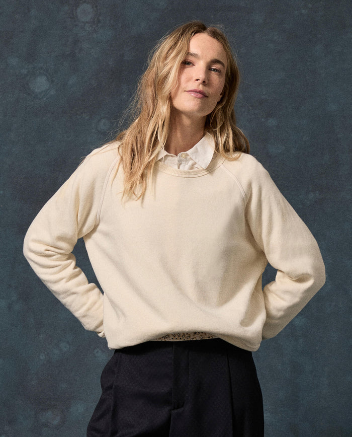 A person with long blond hair wears THE GREAT.’s The Fleece Rookie Sweatshirt in Solid Washed White—crafted in Los Angeles—over a collared shirt and black pants, standing against a dark mottled background.