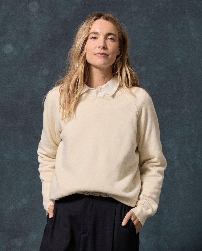 A person with long blond hair, wearing THE GREAT.’s The Fleece Rookie Sweatshirt in Washed White over a white collared shirt and dark pants, stands against a textured dark blue background.