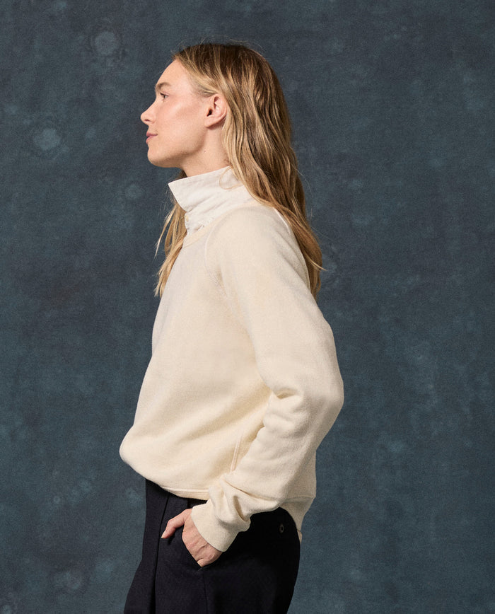 A woman stands in profile against a mottled blue background, wearing THE GREAT.’s The Fleece Rookie Sweatshirt in Solid -- Washed White—a high-collar, elevated vintage sweatshirt from the F26 D1 FLEECE BACK KNITS collection, made in Los Angeles—and dark pants.