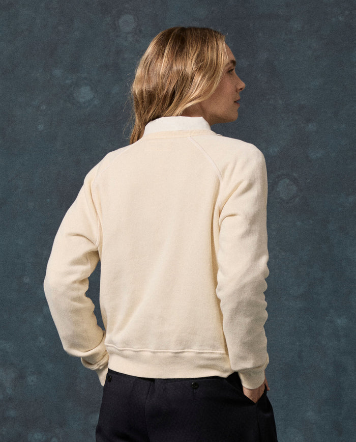 A person with long, light brown hair is seen from behind, wearing THE GREAT.’s The Fleece Rookie Sweatshirt in Solid -- Washed White (F26 D1 FLEECE BACK KNITS), made in Los Angeles, paired with dark pants and standing against a textured, dark background.