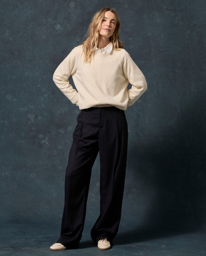 A woman stands against a dark backdrop, wearing THE GREAT. F26 D1 FLEECE BACK KNITS The Fleece Rookie Sweatshirt in Solid -- Washed White—evoking a vintage feel—layered over a collared shirt, paired with wide-leg dark trousers and cream shoes, her hands behind her back.