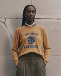 A person stands against a stone wall wearing THE GREAT. F26 D1 The Fleece Rookie Sweatshirt w/ Growling Panther Graphic in mustard, featuring "THE GREAT PANTHERS" and a blue growling panther graphic, paired with khaki pleated pants and a shell necklace.