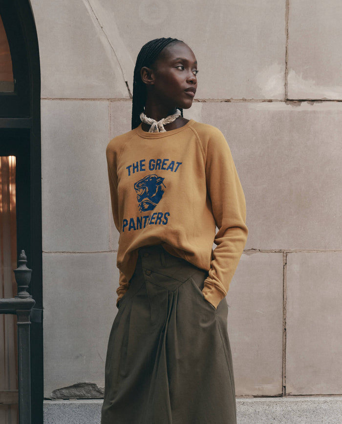 A model wears THE GREAT. F26 D1's The Fleece Rookie Sweatshirt w/ Growling Panther Graphic in mustard, styled with a neck scarf and pleated olive green trousers, posing against a stone-walled building exterior.