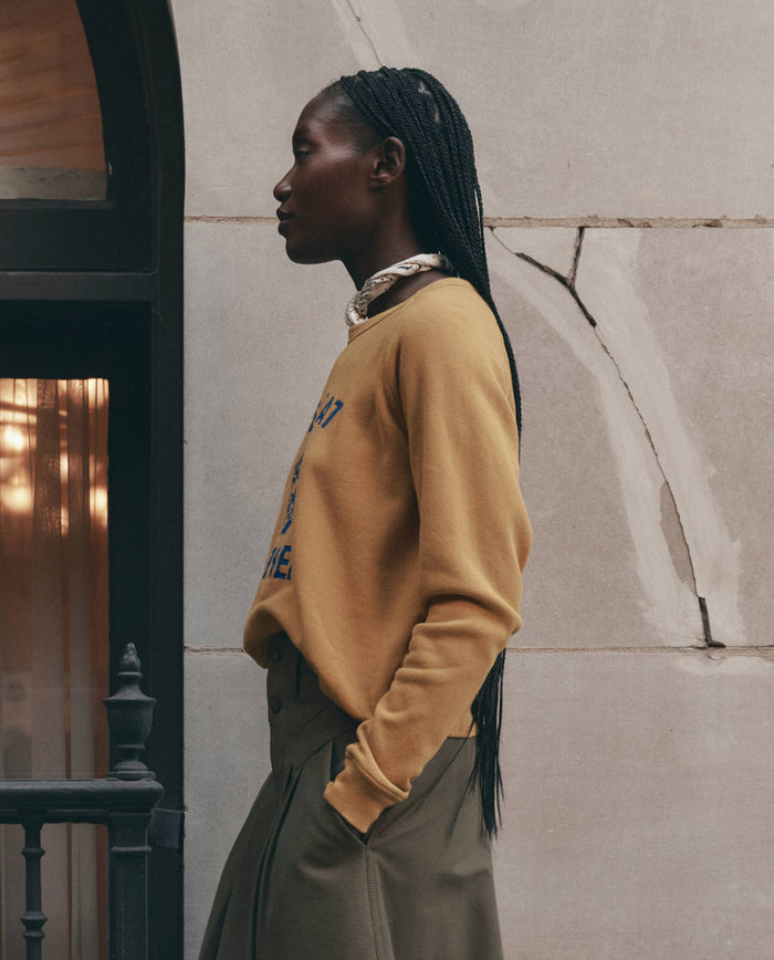 A person with long braids, wearing THE GREAT. F26 D1 The Fleece Rookie Sweatshirt w/ Growling Panther Graphic in mustard and olive pants, stands in profile by a stone wall and window.