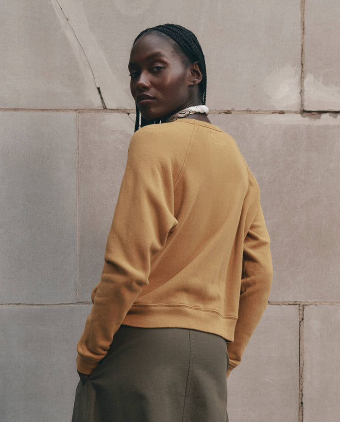 Wearing THE GREAT. F26 D1's The Fleece Rookie Sweatshirt with Growling Panther Graphic in mustard and an olive skirt, a person with braided hair stands before a stone wall, glancing over their shoulder like a prowling panther.
