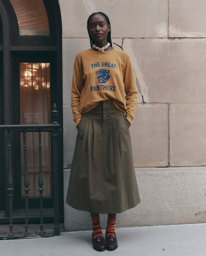 A person stands against a stone wall wearing THE GREAT. F26 D1 The Fleece Rookie Sweatshirt w/ Growling Panther Graphic in mustard, paired with a long olive-green pleated skirt, striped socks, and brown loafers.