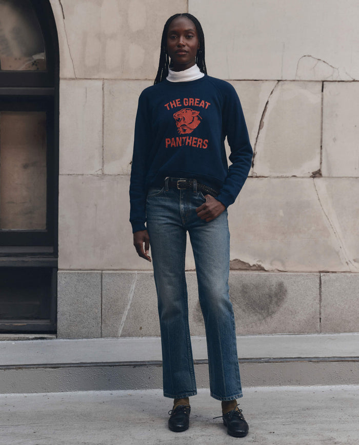 A person stands outside against a worn stone wall, wearing THE GREAT. F26 D1 The Fleece Rookie Sweatshirt w/ Growling Panther Graphic in True Navy, paired with jeans, black shoes, and a white turtleneck.