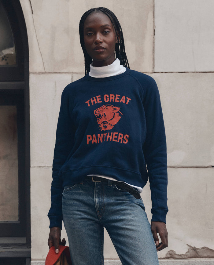 A person is standing outdoors, dressed in THE GREAT. F26 D1 The Fleece Rookie Sweatshirt w/ Growling Panther Graphic in True Navy, paired with blue jeans and a white turtleneck.