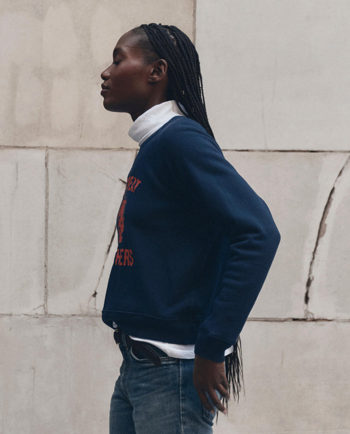 A person with long braids stands in profile against a light-colored stone wall, wearing THE GREAT.'s The Fleece Rookie Sweatshirt w/ Growling Panther Graphic in True Navy over a white turtleneck and blue jeans.