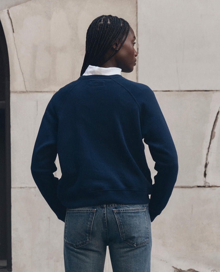 A person with braided hair stands facing away against a light stone wall, wearing THE GREAT.’s The Fleece Rookie Sweatshirt w/ Growling Panther Graphic in True Navy, layered over a white collared shirt and blue jeans.