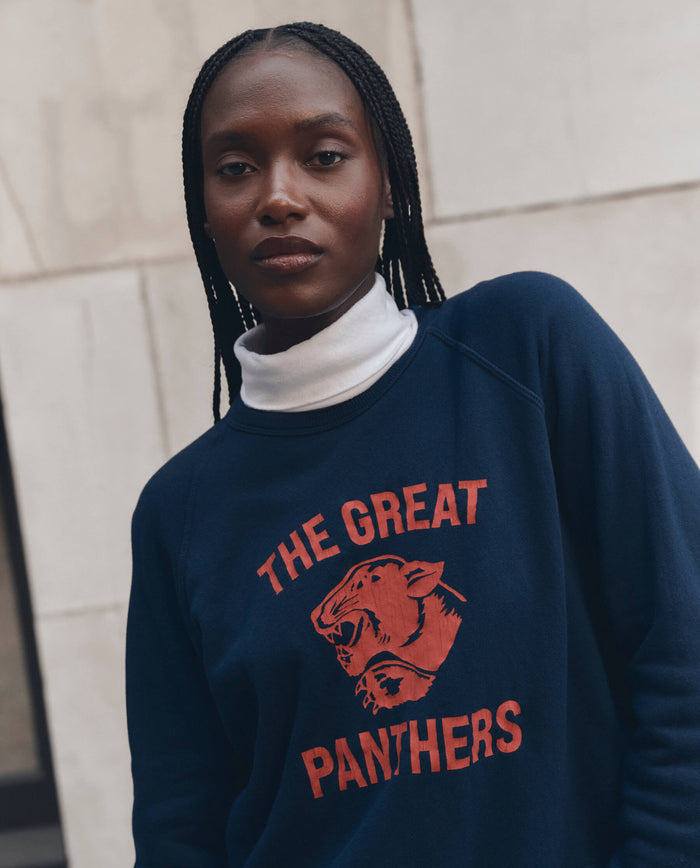 A person with braided hair stands in front of a stone wall, wearing THE GREAT. F26 D1 The Fleece Rookie Sweatshirt w/ Growling Panther Graphic in True Navy over a white turtleneck.