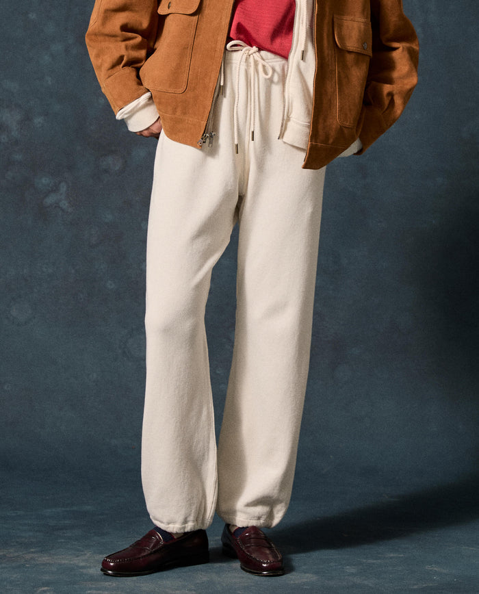 Wearing THE GREAT. F26 D1 FLEECE BACK KNITS The Fleece Sport Sweatpant in Washed White, a person stands against a dark background, paired with a red shirt, brown jacket, and dark loafers.