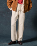 Wearing THE GREAT. F26 D1 FLEECE BACK KNITS The Fleece Sport Sweatpant in Washed White, a person stands against a dark background, paired with a red shirt, brown jacket, and dark loafers.