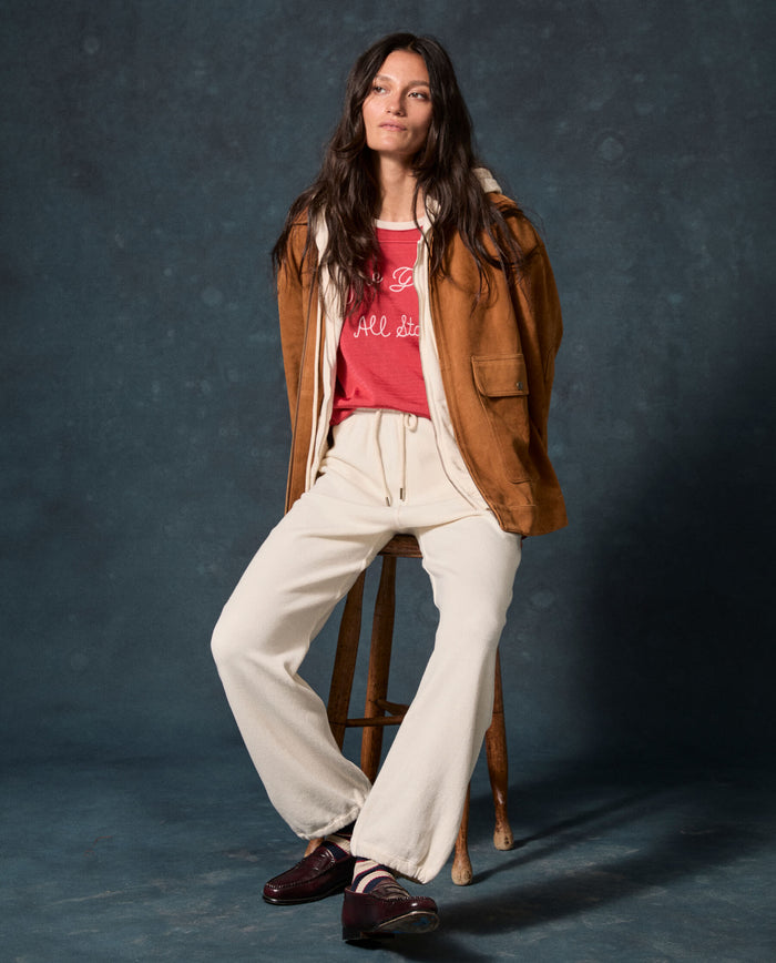 A woman sits on a stool against a dark studio backdrop, wearing a brown jacket, red shirt, dark loafers, and THE GREAT. F26 D1 FLEECE BACK KNITS The Fleece Sport Sweatpant in Washed White.