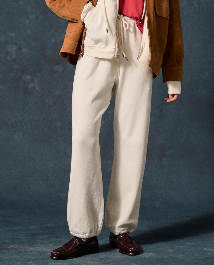 Wearing THE GREAT. F26 D1 FLEECE BACK KNITS The Fleece Sport Sweatpant in Washed White, paired with a red shirt, cream hoodie, brown jacket, and dark loafers, this individual stands against a dark textured background.