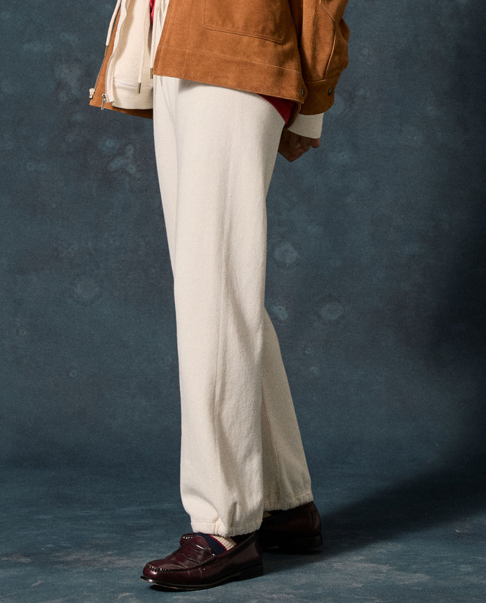 A person wearing THE GREAT. F26 D1 FLEECE BACK KNITS ‘The Fleece Sport Sweatpant’ in Washed White is shown from the waist down, paired with a brown jacket and dark loafers, standing against a textured dark blue background.