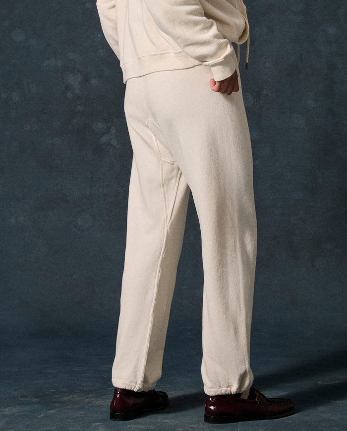 A person stands with one hand in their pocket, wearing THE GREAT. F26 D1 FLEECE BACK KNITS “The Fleece Sport Sweatpant” in Washed White and a matching sweatshirt, paired with dark brown dress shoes against a dark, textured background.