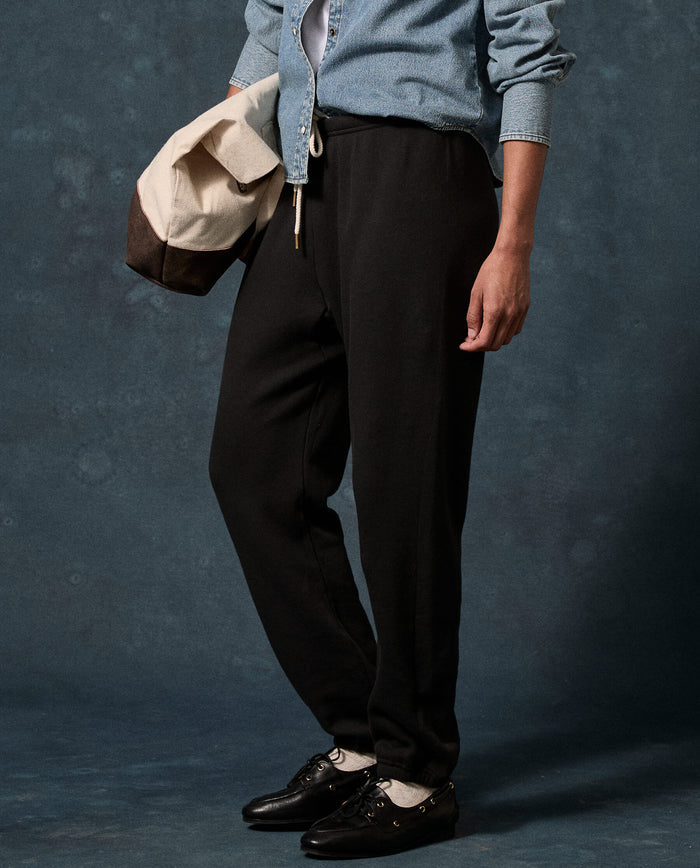 A person wearing a denim shirt, The Fleece Stadium Sweatpant in Solid -- Almost Black by THE GREAT. F26 D1 FLEECE BACK KNITS, and black shoes stands against a dark background, holding a beige and brown bag.