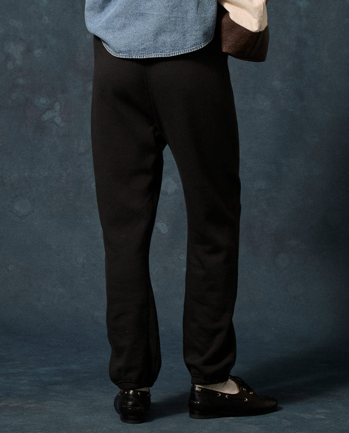 A person is standing on a blue floor, photographed from behind at full leg length, wearing THE GREAT. F26 D1 FLEECE BACK KNITS The Fleece Stadium Sweatpant in Solid -- Almost Black, paired with black shoes and a blue and white top.