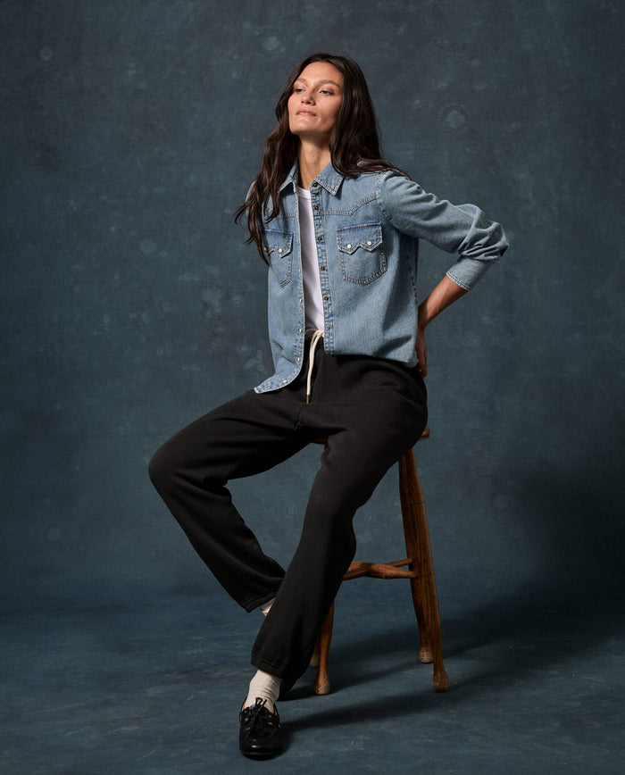 A woman sits on a wooden stool against a dark backdrop, wearing a denim shirt over a white top, and THE GREAT. F26 D1 FLEECE BACK KNITS The Fleece Stadium Sweatpant in Solid -- Almost Black, paired with white socks and black shoes.