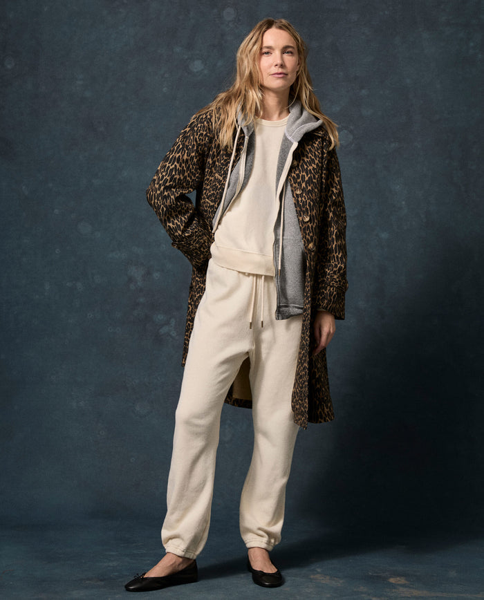 A person stands against a dark mottled background wearing THE GREAT. F26 D1 FLEECE BACK KNITS The Fleece Stadium Sweatpant in Washed White, paired with a gray hoodie, a leopard print coat, and black flats.