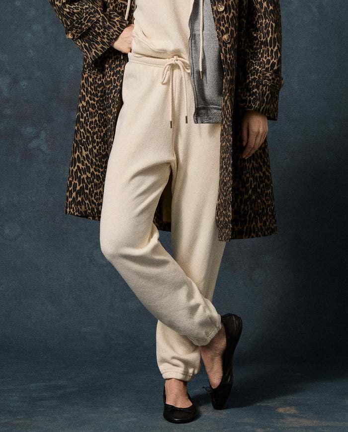 A person standing with legs crossed against a dark background, wearing THE GREAT. F26 D1 FLEECE BACK KNITS “The Fleece Stadium Sweatpant” in Solid -- Washed White, paired with a light top, grey hoodie, leopard print coat, and black ballet flats.