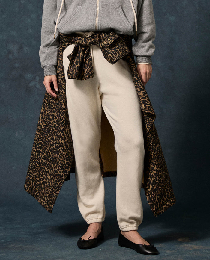 A person stands against a dark background wearing a grey zip-up hoodie, black flats, and THE GREAT. F26 D1 FLEECE BACK KNITS The Fleece Stadium Sweatpant in Solid Washed White, with a leopard print coat tied around the waist.