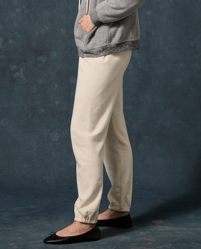 A person wearing a gray hoodie, THE GREAT. The Fleece Stadium Sweatpant in Washed White, and black ballet flats stands against a dark blue textured background.