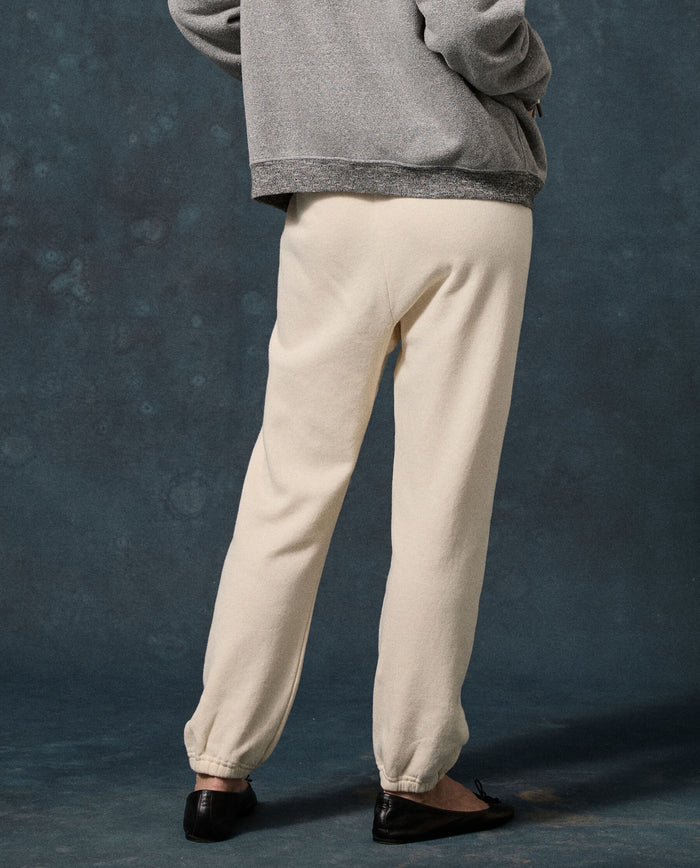 Wearing THE GREAT. The Fleece Stadium Sweatpant in Washed White, a person stands with their back to the camera against a dark textured background.