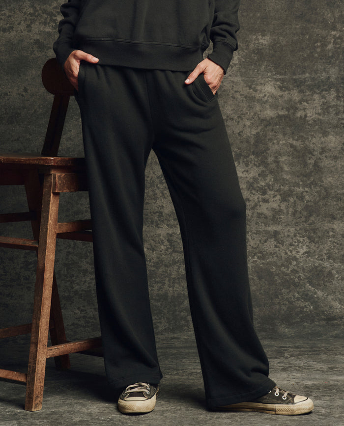 A person stands with hands in pockets wearing THE GREAT. H25 CORE FLEECEBACK The Fleece Victory Sweatpant in Almost Black, paired with a black sweatshirt and gray sneakers beside a wooden chair against a textured gray backdrop.