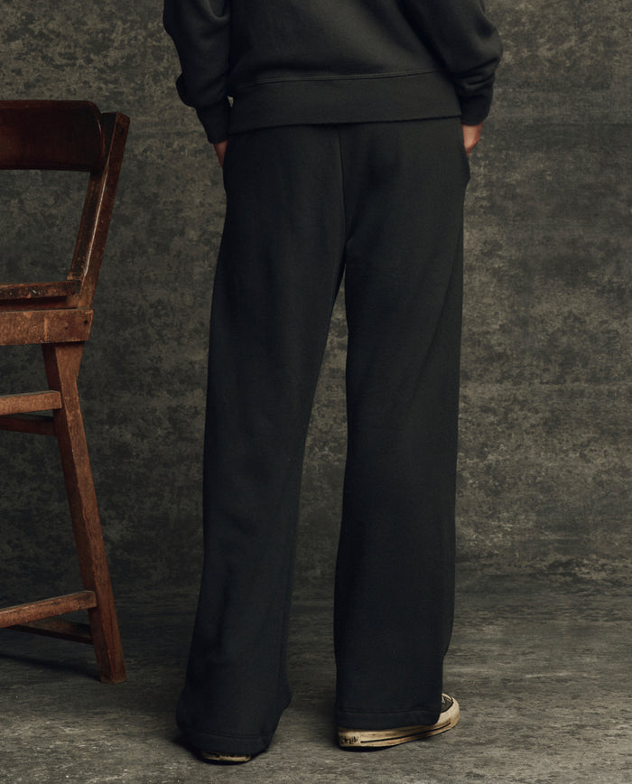 Someone wearing THE GREAT. H25 CORE FLEECEBACK The Fleece Victory Sweatpant in Almost Black and sneakers stands with their back to the camera beside a wooden chair, set against a textured gray backdrop.