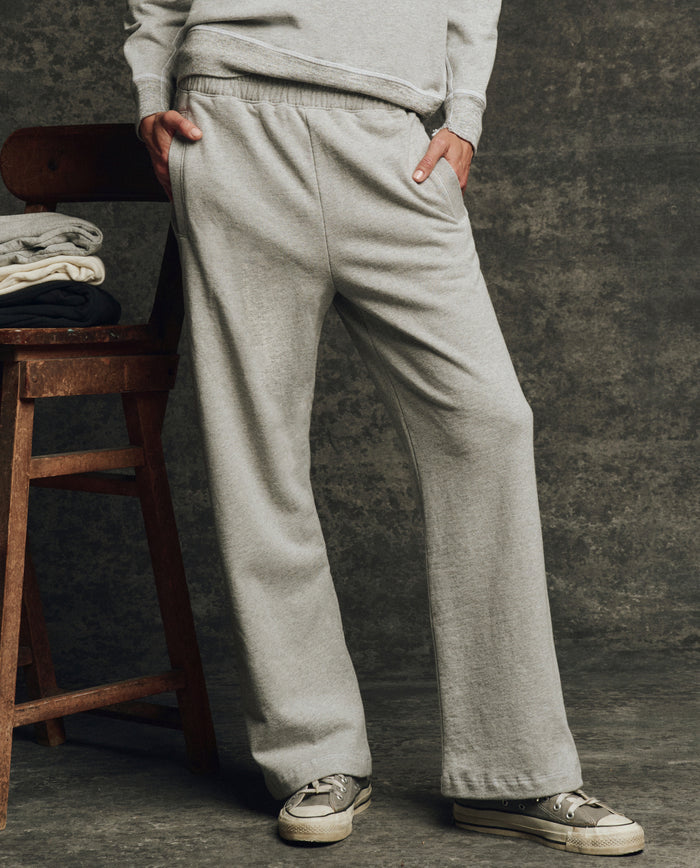 A person wears THE GREAT. H25 CORE FLEECEBACK The Fleece Victory Sweatpant in Heather Grey with sneakers, standing by a wooden chair stacked with folded clothes against a dark textured background.