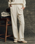 Wearing THE GREAT. H25 CORE FLEECEBACK The Fleece Victory Sweatpant in Washed White, a person stands with hands in pockets by a wooden chair stacked with folded clothes, set against a textured gray backdrop.