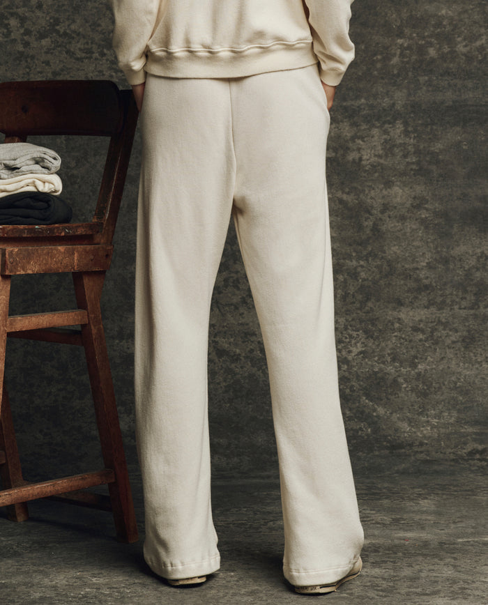 A person stands with hands in pockets, wearing THE GREAT. H25 CORE FLEECEBACK The Fleece Victory Sweatpant in Washed White and a matching sweatshirt, facing away. A wooden chair with folded clothes is on the left.