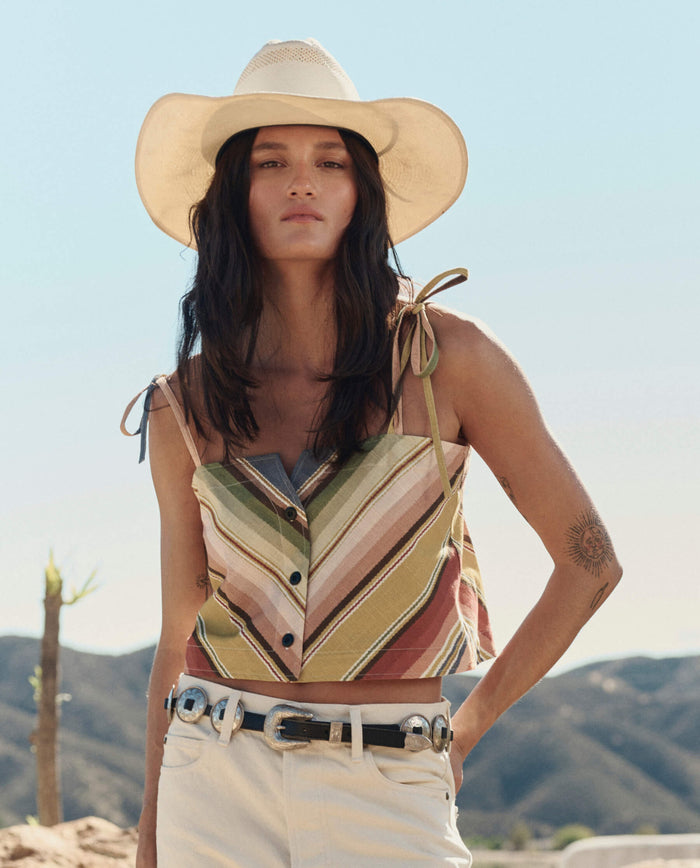 A person wearing THE GREAT. PF26’s The Folio Tank in Davenport Stripe, paired with a wide-brimmed hat and belted white pants, stands outdoors with hills and a clear sky in the background.