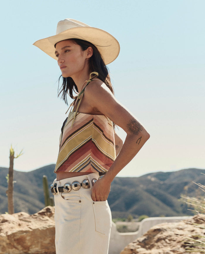 Wearing THE GREAT. PF26’s The Folio Tank in Davenport Stripe, a person with a wide-brimmed hat, white pants, decorative belt, and arm tattoo stands outdoors against mountains, stylish leather folio in hand.