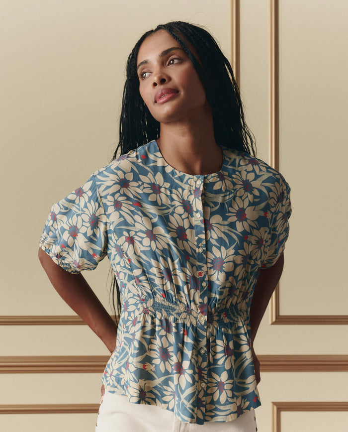 A woman with long braided hair wears THE GREAT. SP26 D2 Hayflower Top in Blue Scattered Bloom, a short-sleeve floral blouse with buttons, paired with white pants, standing against a beige paneled wall.
