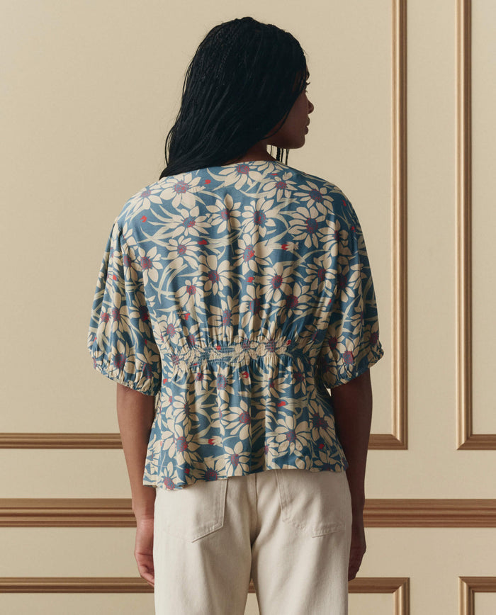 Person stands with back to camera, wearing THE GREAT. SP26 D2 Hayflower Top in Blue Scattered Bloom (floral, short sleeves), paired with light pants against a beige paneled wall.
