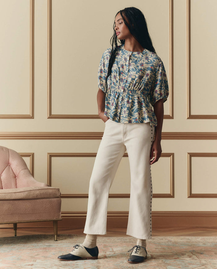 A woman stands indoors wearing a floral blouse, The Studded Wayne Jean in Natural by THE GREAT. SP26 D2, and two-tone shoes, beside a pink upholstered chair and a wall with decorative molding.