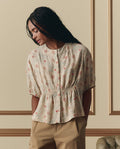 A woman with long braided hair wears THE GREAT.'s Hayflower Top in Champagne Scattered Bloom, standing indoors against a decorative wall and pairing it with beige pants.