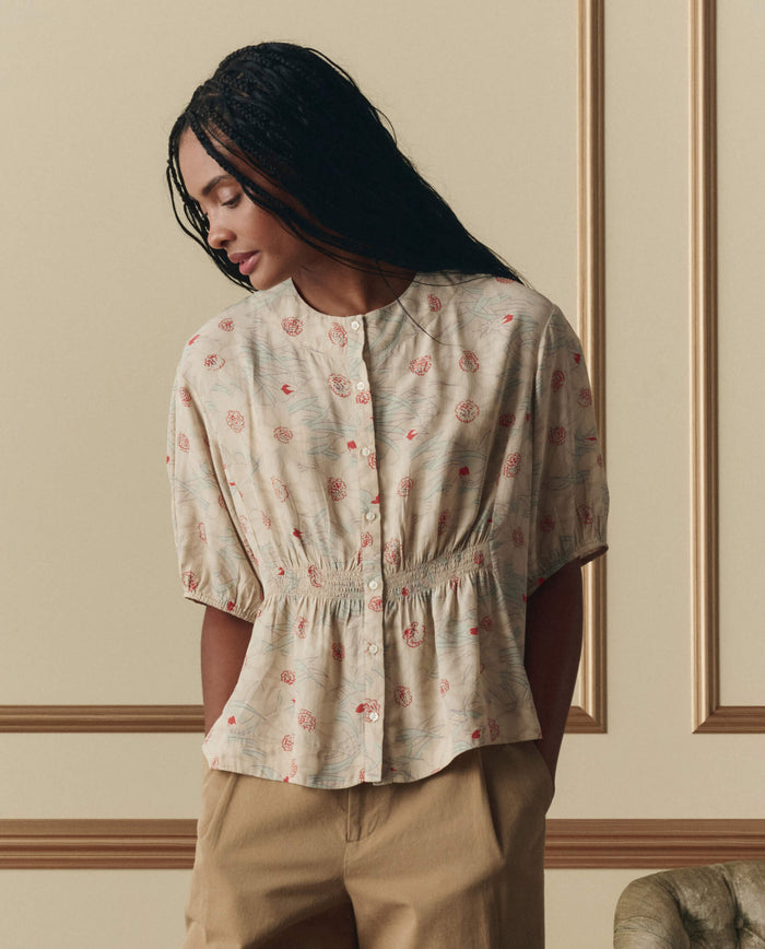 A woman with long braided hair wears THE GREAT.'s Hayflower Top in Champagne Scattered Bloom, standing indoors against a decorative wall and pairing it with beige pants.