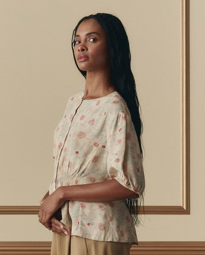 A woman with long braided hair stands before a beige wall, wearing THE GREAT. SP26 D2's The Hayflower Top in Champagne Scattered Bloom and tan pants.