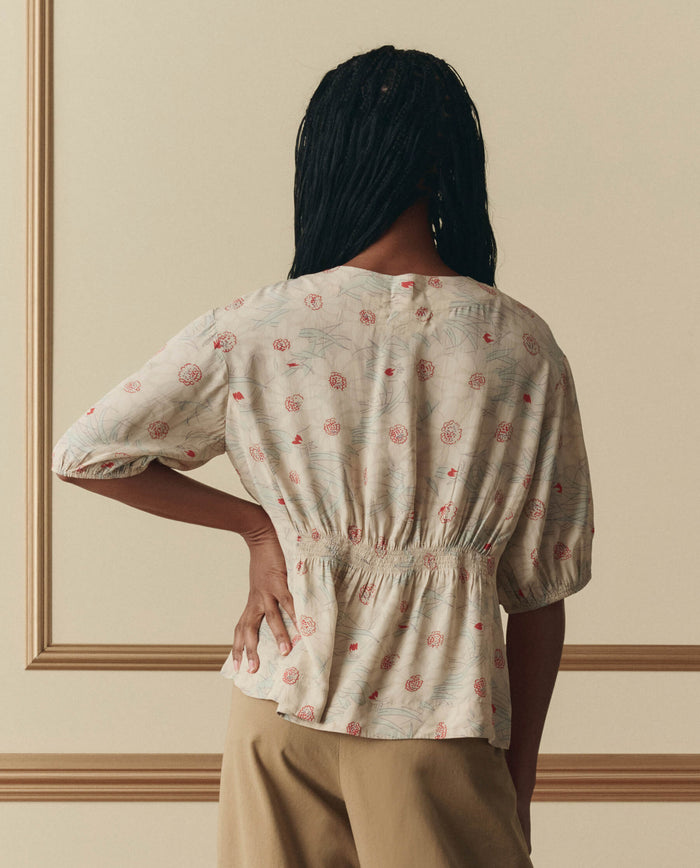 A person stands facing away, wearing THE GREAT.'s Hayflower Top in Champagne Scattered Bloom— a light floral short-sleeve blouse— paired with tan pants, against a neutral background with decorative molding.